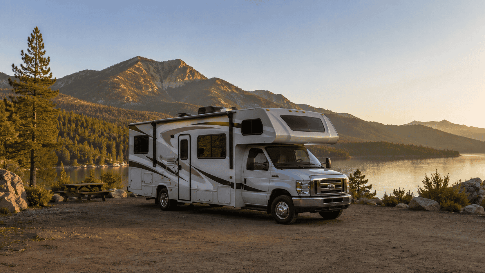 Class C motorhome parked at a lakeside campsite at sunset with forested mountains in the background.