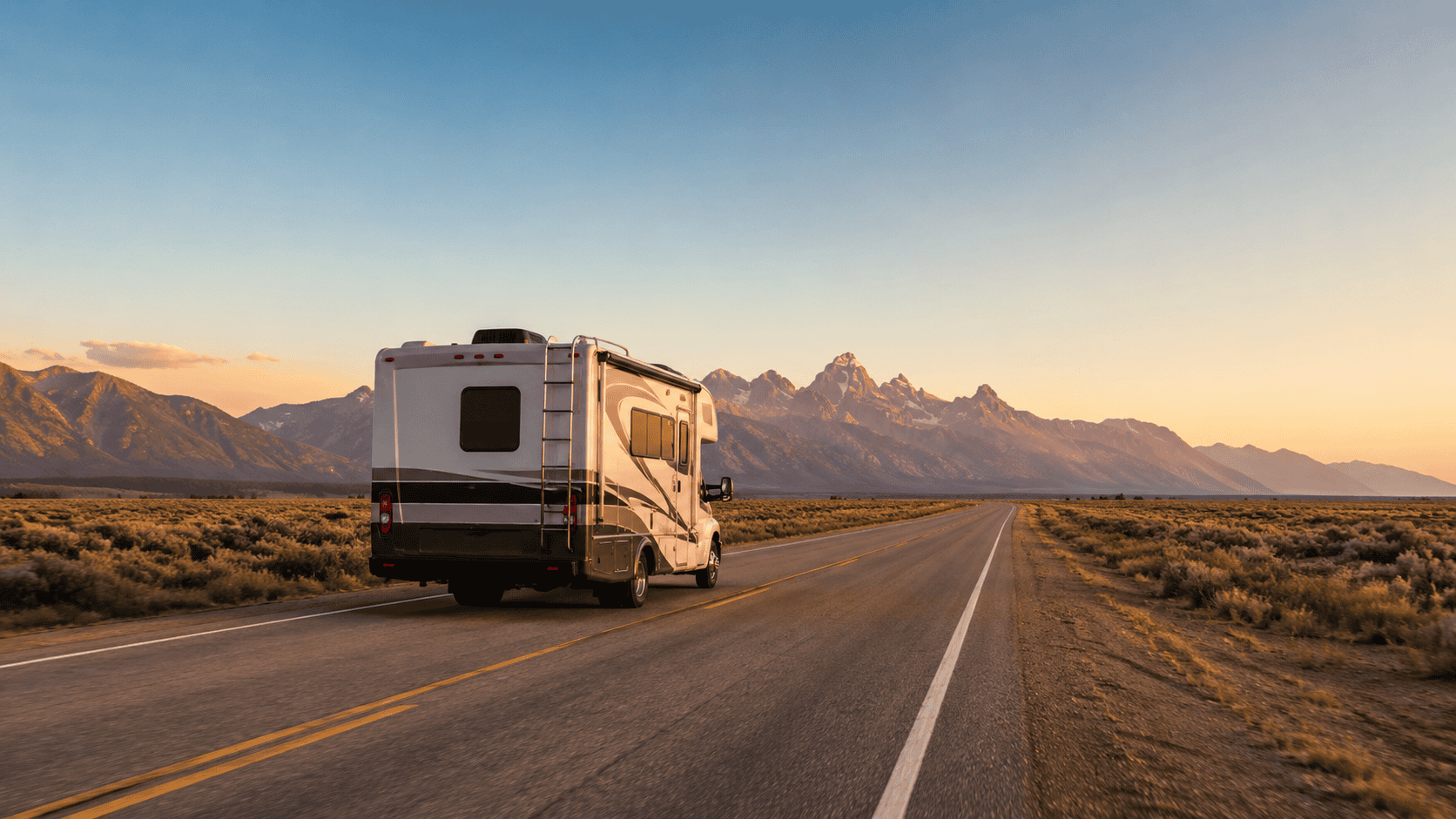 Class C motorhome on an open highway heading toward distant mountains at sunset.