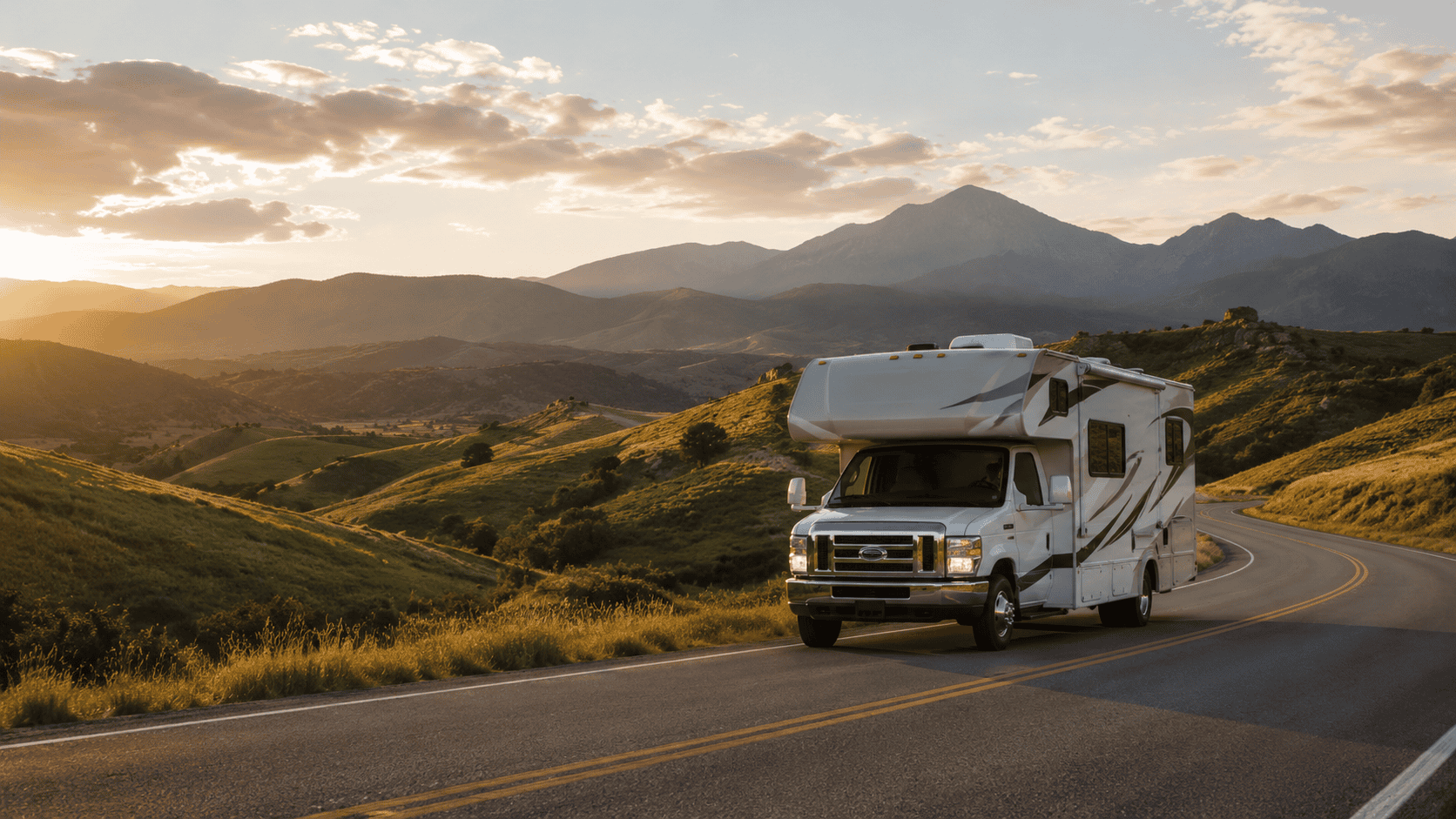 Modern Class C motorhome driving along a winding highway through green rolling hills toward distant mountains at golden hour.